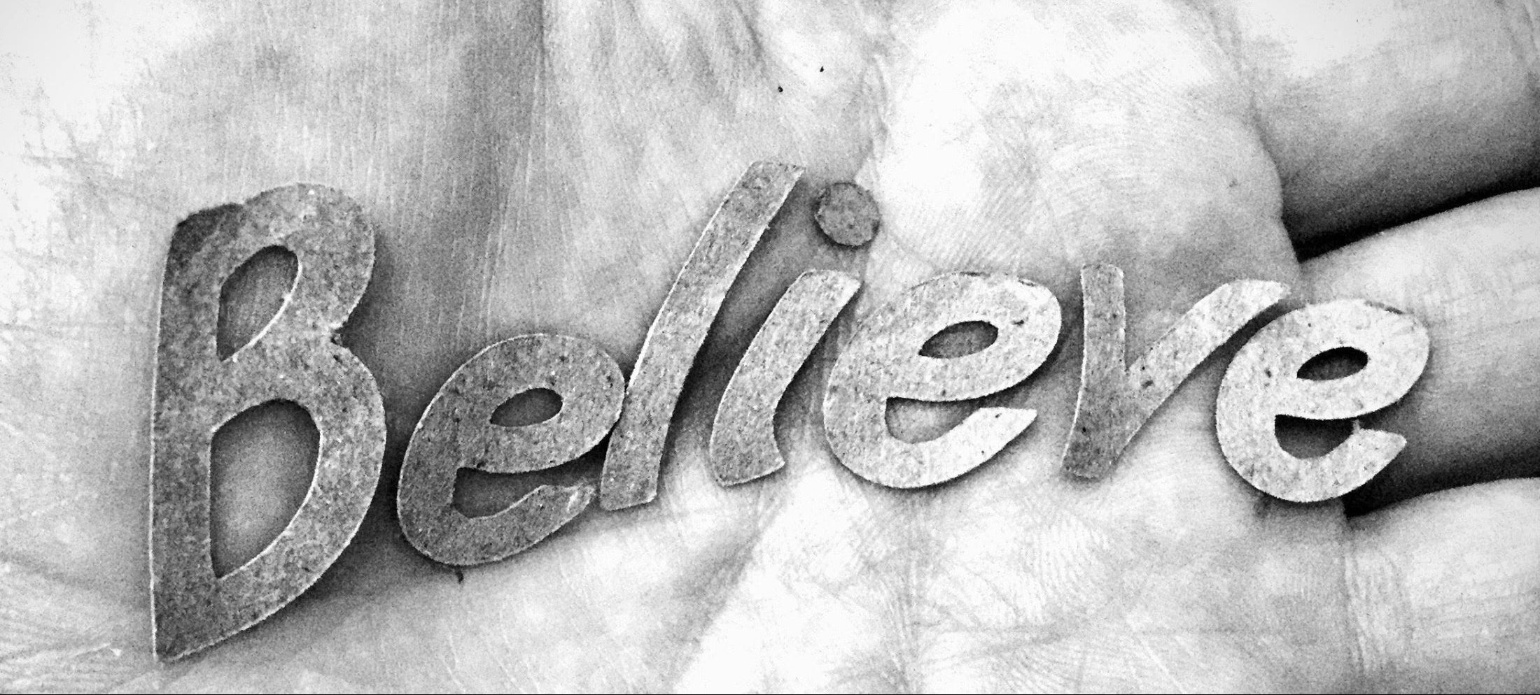 The word 'Believe' spelled out in 3D metallic letters resting on a textured human hand, shown in black and white.